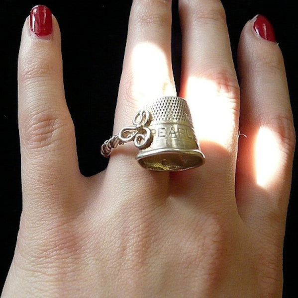 I'm all about the stories behind things worn or imperfect, for they speak of a life well-lived. A copy of a dented and lovingly used old thimble engraved with "Pearls" and resting above a twisted wire shank, accented by a little braided "eye"missing its "hook"  1--1/2" x 7/8" Size 4-10 approximately. Made in the USA. Hand signed.  Sterling Silver Dented Thimble Ring